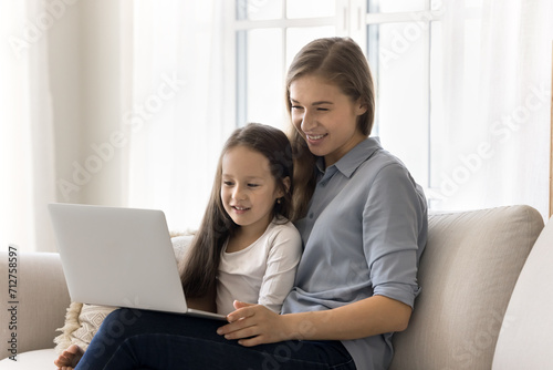 Mother and little daughter spend weekend leisure on internet use laptop, sit on sofa look at device screen, watch educational program, buy in marketplace, enjoy e-commerce services and cartoons online