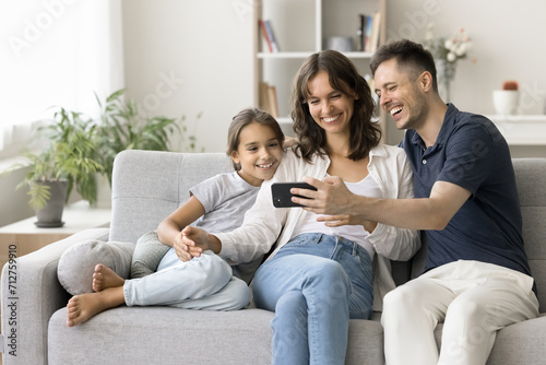 Happy young parents and cute daughter kid watching content on smartphone, using gadget for online communication, taking family selfie on cozy home couch, enjoying Internet technology