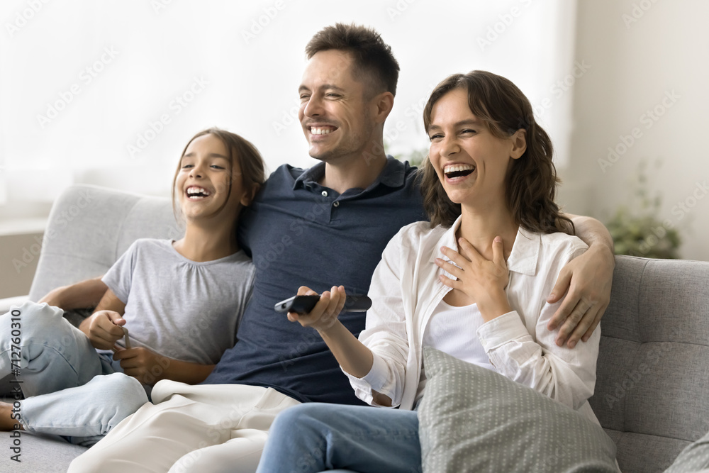 Cheerful excited young mom, dad and daughter kid watching TV at home together, relaxing on couch, using distant remote control, looking away, laughing, enjoying movie, series, Internet television Cheerful excited young mom, dad and daughter kid watching TV at home together, relaxing on couch, using distant remote control, looking away, laughing, enjoying movie, series, Internet television
