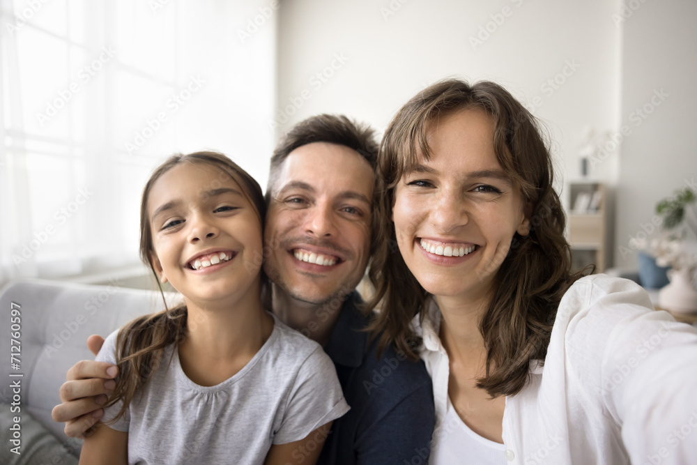 Happy young parents and cute girl kid looking at camera with toothy smiles, posing for home family selfie. Cheerful mother holding gadget with camera, taking self portrait with husband and daughter Happy young parents and cute girl kid looking at camera with toothy smiles, posing for home family selfie. Cheerful mother holding gadget with camera, taking self portrait with husband and daughter