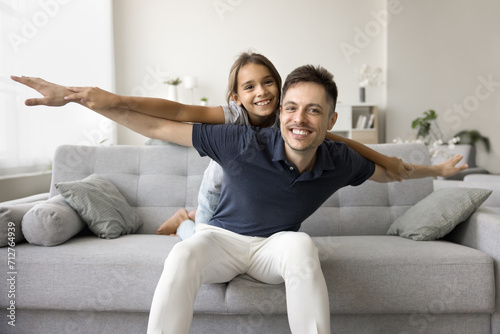 Cheerful playful daddy piggybacking little daughter kid with open flying hands. Adorable girl playing airplane on fathers back, enjoying activity, funny leisure, playtime on home sofa. Family portrait photo