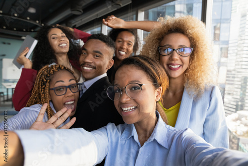 Office selfie. Friendly young biracial workers colleagues have fun at workplace shoot cute silly self picture on phone. Active millennial black business team take funny group photo for corporate blog