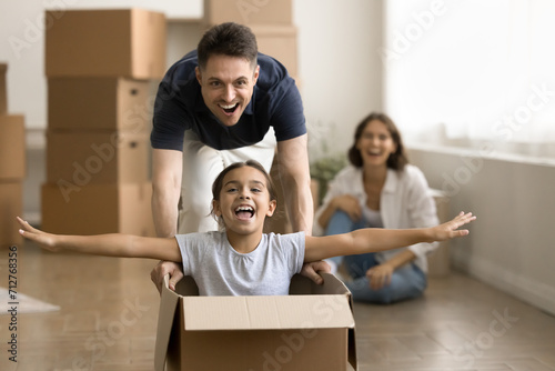 Excited strong young dad walking and driving relocation box with cheerful kid inside, playing with daughter. Family celebrating apartment, house buying, having fun among paper containers photo