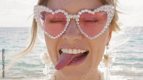 Portrait of funny hilarious woman in pink glasses making silly faces, pretending singing outloud on beach. Positive girl having good time showing tongue and playing kissy-face. Closeup emotional bride
