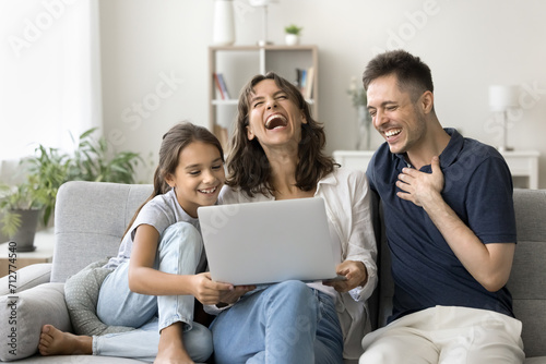 Joyful excited family couple and cute little kid using laptop at home, promoting online communication, domestic Internet technology, wireless connection, resting on sofa with computer, laughing