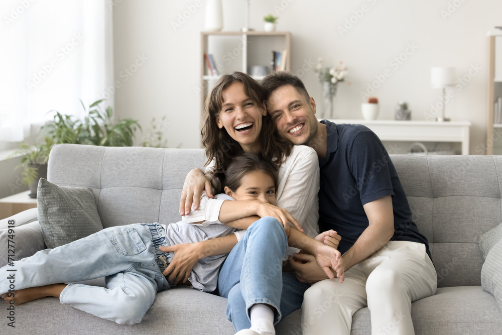 Cheerful young couple of parents tightening sweet child girl on cozy sofa, enjoying family leisure in stylish comfortable home interior, smiling, laughing, looking at camera for portrait
