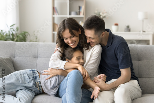 Happy young parents cuddling sweet adorable kid girl on comfortable home couch, hugging child with love, adoration, laughing, spending family leisure with daughter child together