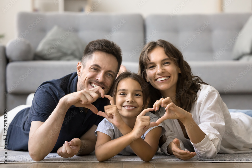 Cheerful attractive mom, dad and sweet daughter kid joining fingers, showing hand heart shapes, resting together on warm floor at cozy home, looking at camera with toothy smiles for family portrait Cheerful attractive mom, dad and sweet daughter kid joining fingers, showing hand heart shapes, resting together on warm floor at cozy home, looking at camera with toothy smiles for family portrait