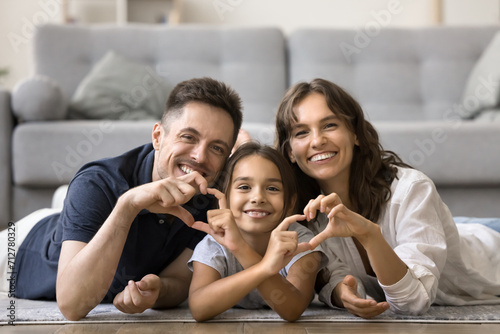 Cheerful attractive mom, dad and sweet daughter kid joining fingers, showing hand heart shapes, resting together on warm floor at cozy home, looking at camera with toothy smiles for family portrait © fizkes