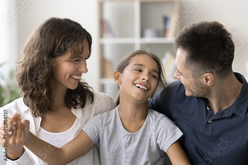 Cheerful young parents and happy carefree child girl sitting close together  talking  laughing  hugging  enjoying family leisure  relationships  childhood  parenthood. Mom and dad cuddling kid