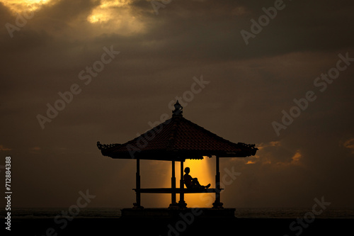 A solitary figure contemplates the expanse of cloudy sky from a peaceful gazebo, a warm shaft of sunlight casting a tranquil silhouette against serene waters below