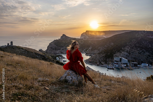 Woman sunset sea mountains. Happy woman siting with her back on the sunset in nature summer posing with mountains on sunset, silhouette. Woman in the mountains red dress, eco friendly