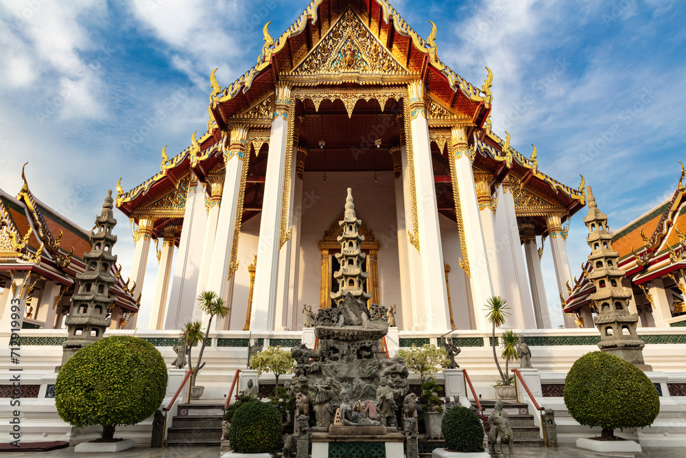 Entrance to Wat Suthat Temple in Bangkok, thailand. Ornamental garden in front. Other buildings on either side; blue sky and clouds above.
Entrance to Wat Suthat Temple in Bangkok, thailand. Ornamental garden in front. Other buildings on either side; blue sky and clouds above.