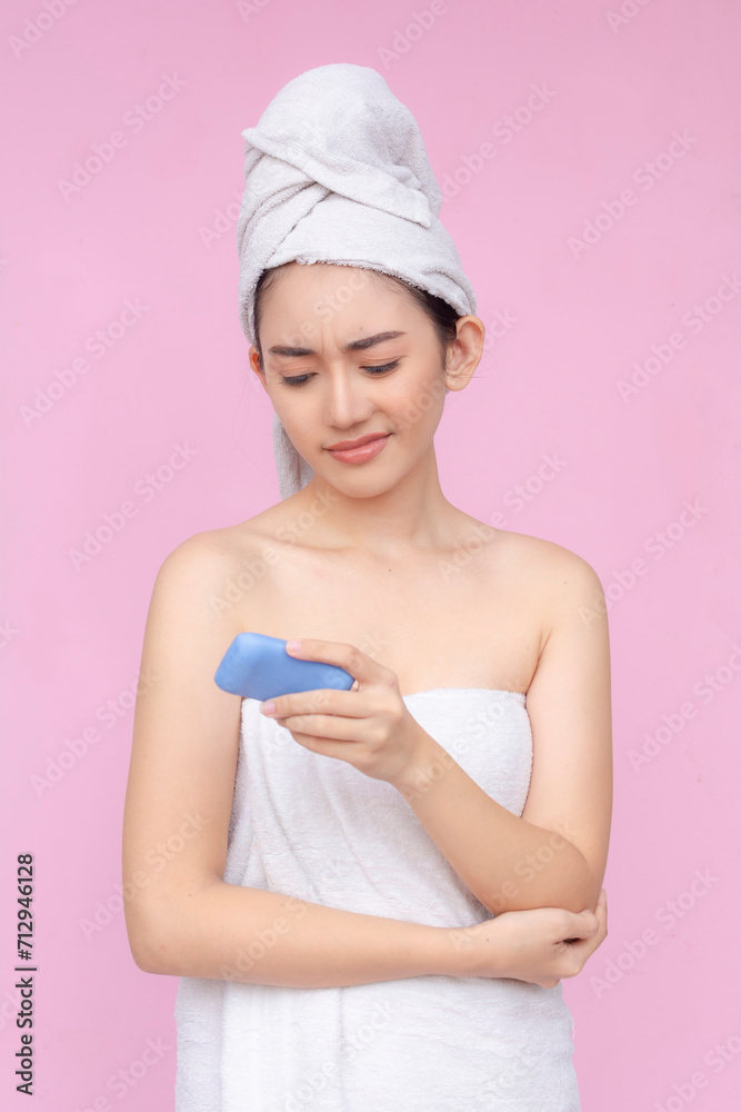 A young asian woman wrapped in a towel hesitant to use a blue deodorant body soap. Bath and body care concept. Soft pInk background. A young asian woman wrapped in a towel hesitant to use a blue deodorant body soap. Bath and body care concept. Soft pInk background.