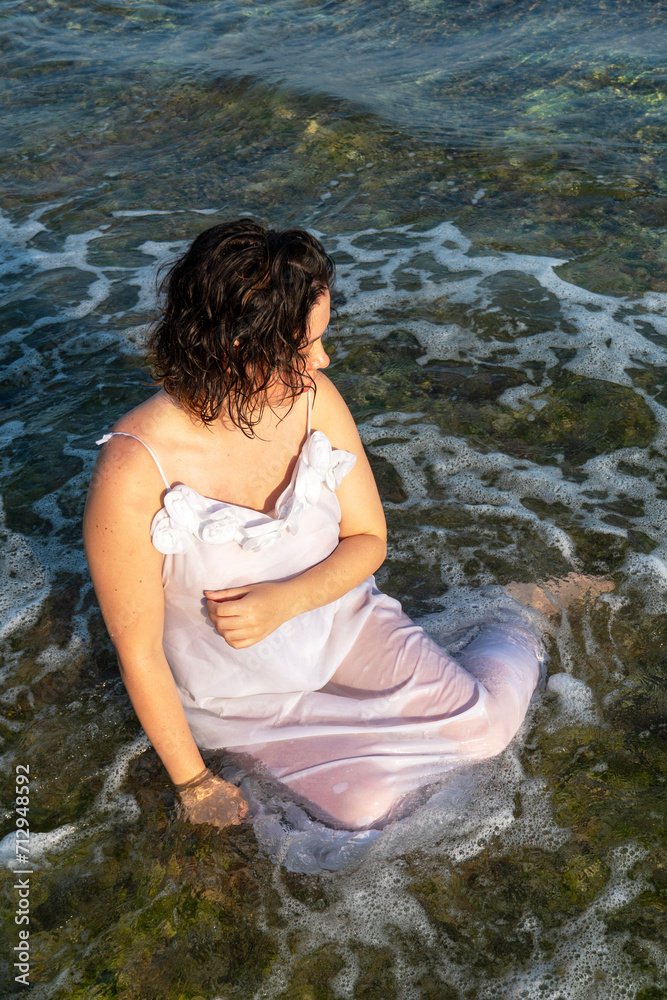 Beautiful woman in white dress sits in the water on ocean shores