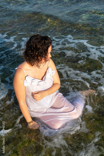 Beautiful woman in white dress sits in the water on ocean shores © Fruityk