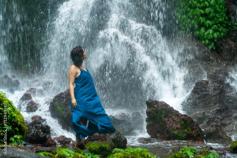 Portrait of the young beautiful woman standing in front of a waterfall in an blue dress.