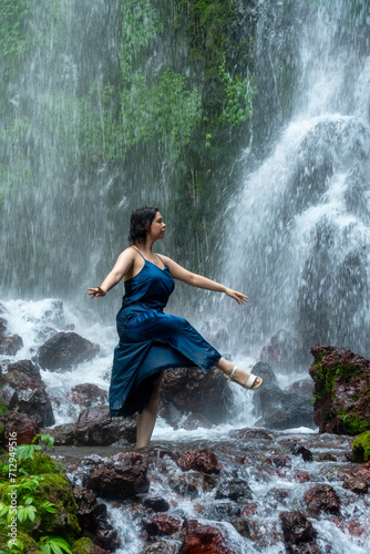 Portrait of the young beautiful woman standing in front of a waterfall in an blue dress.