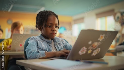 Portrait of a Cute Little African Boy with Stylish Hair Sitting Behind a Desk in Class in Elementary School. Young Pupil Using a Laptop Computer while Listening to a Teacher with Other Kids photo