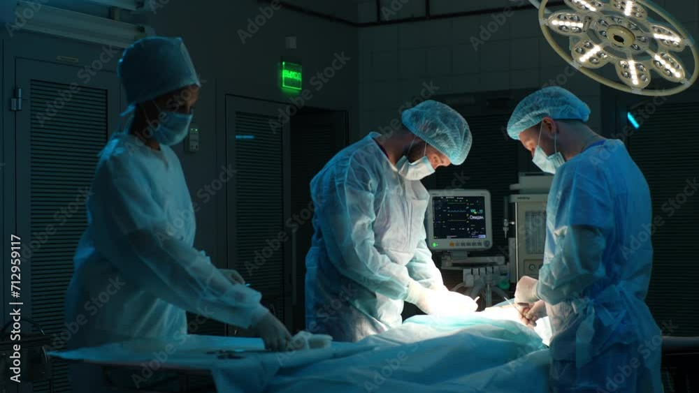 Diverse cooperating team of doctors and surgeons processing surgical operation in dark operating room modern hospital emergency department. Doctors wearing sterile gloves, surgical uniforms and masks. Diverse cooperating team of doctors and surgeons processing surgical operation in dark operating room modern hospital emergency department. Doctors wearing sterile gloves, surgical uniforms and masks.