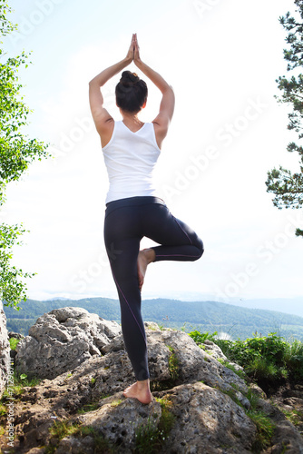 Attractive young woman doing a yoga pose for balance and stretching staying on top of high rock in the mountains