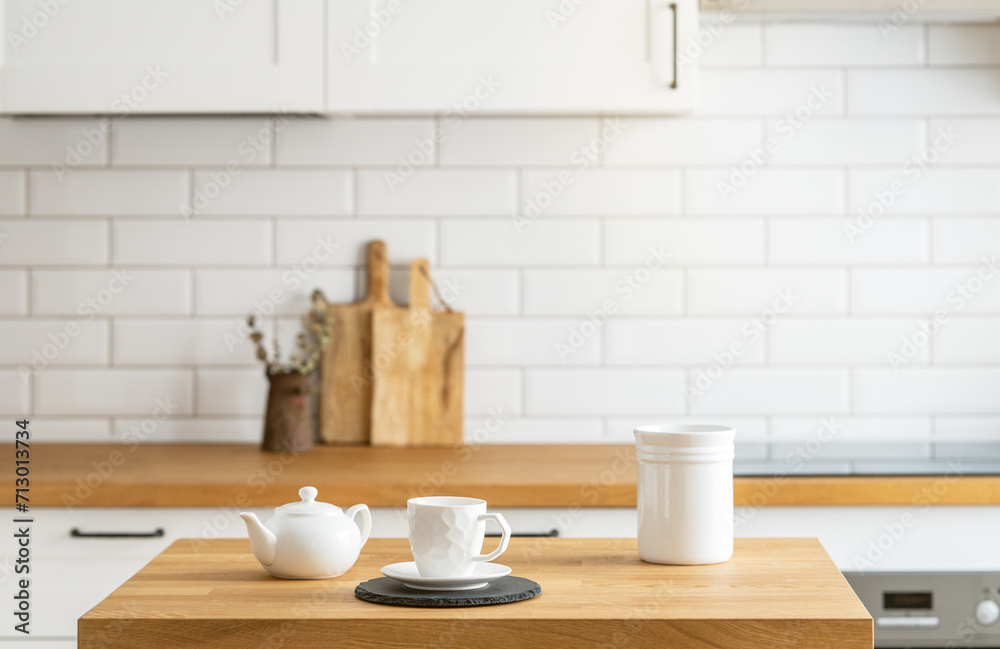 Wooden oak table with a cup of tea and a kettle  in front of the kitchen with a white brick background.