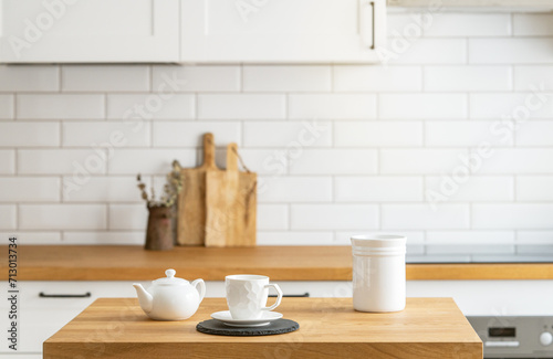 Wooden oak table with a cup of tea and a kettle in front of the kitchen with a white brick background.