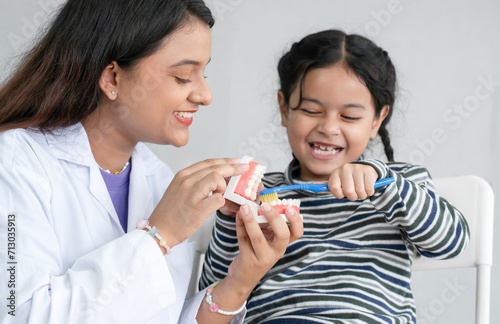 Cute Asian little girl learning how to brush her teeth and gums on a jaw model from young Indian dentist with toothbrush at clinic. Oral and Dental Health. Selective focus on dentist photo