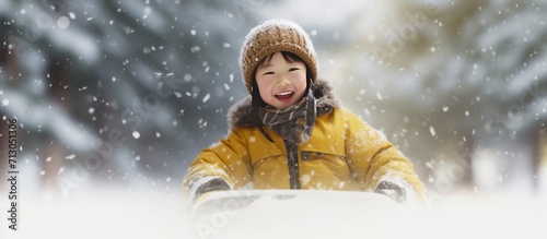 Happy Asian child in yellow down coat sledding in winter down a hill, on blurred winter forest or skiing resort background, with copy space, concept of family winter