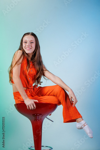 A girl in an orange suit and with a smile on her face poses while sitting on a chair