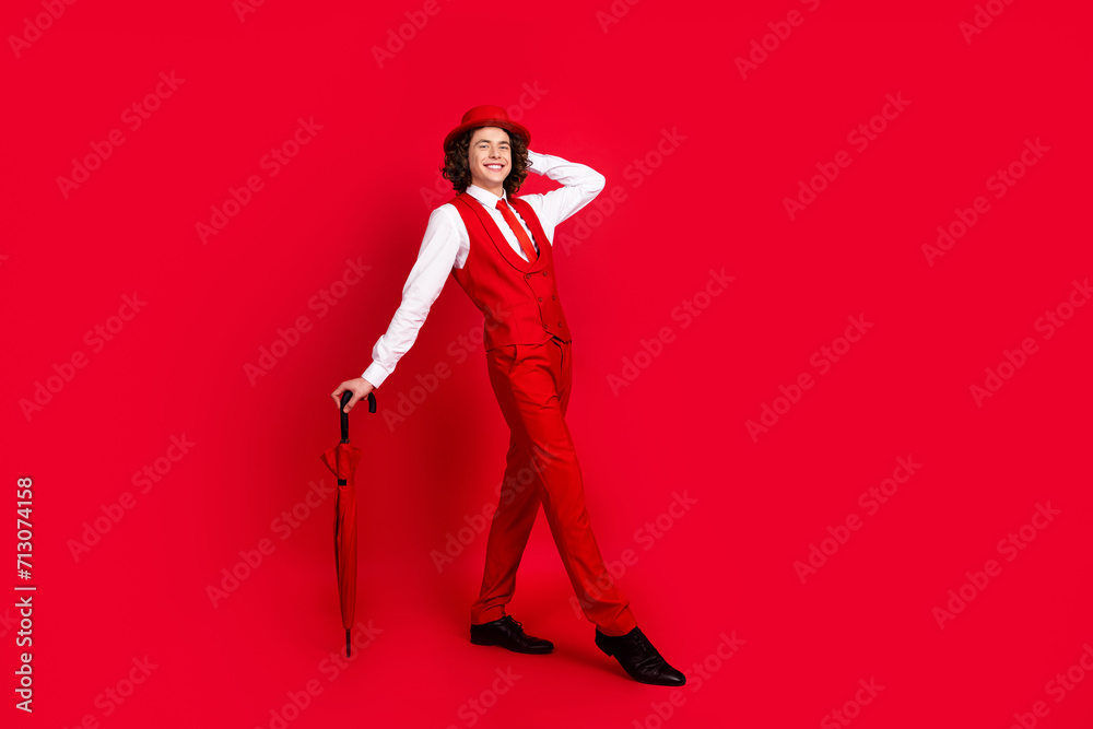 Full body length photo of luxury model teenager guy in suit vest with parasol touching hat showman concert isolated on red color background Full body length photo of luxury model teenager guy in suit vest with parasol touching hat showman concert isolated on red color background