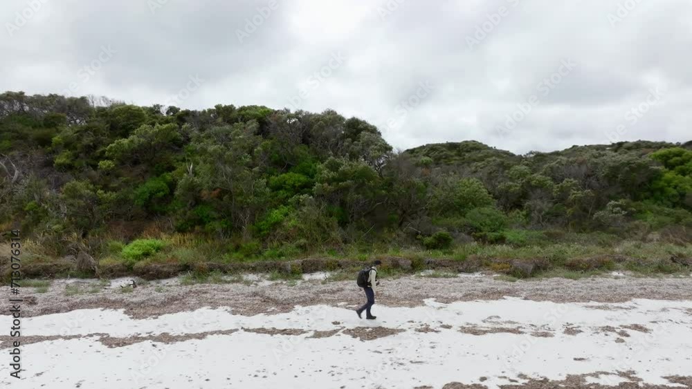 Drone circles around from side on to in front of a man walking along the beach, revealing what is behind him. Drone circles around from side on to in front of a man walking along the beach, revealing what is behind him.