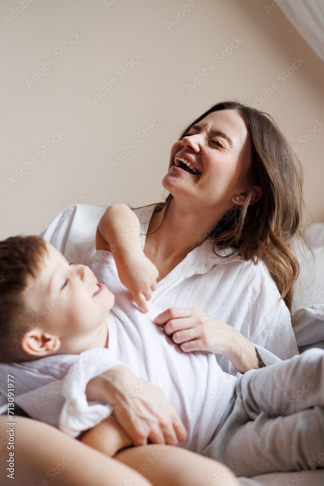 Cheerful woman in shirt hugging toddler son on bed in morning Cheerful woman in shirt hugging toddler son on bed in morning