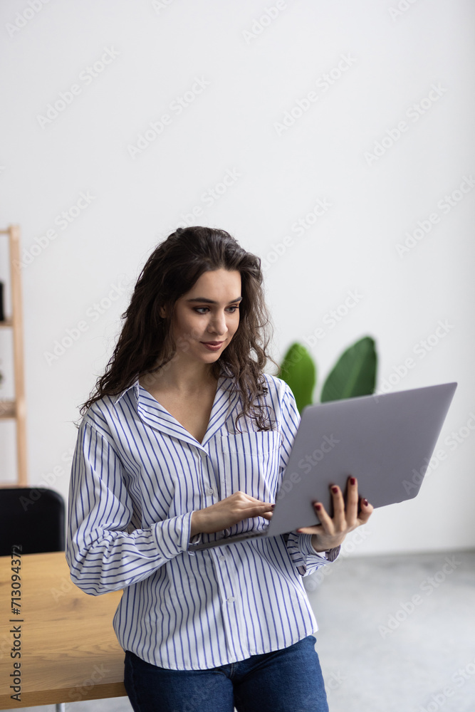Beautiful young business woman wearing shirt using laptop while standing in modern workspace Beautiful young business woman wearing shirt using laptop while standing in modern workspace