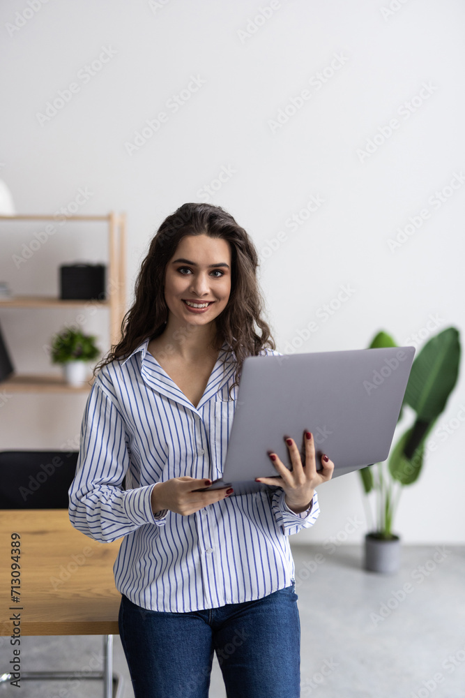 Smiling business woman is standing in the office by the window with a laptop in her hands. Smiling business woman is standing in the office by the window with a laptop in her hands.