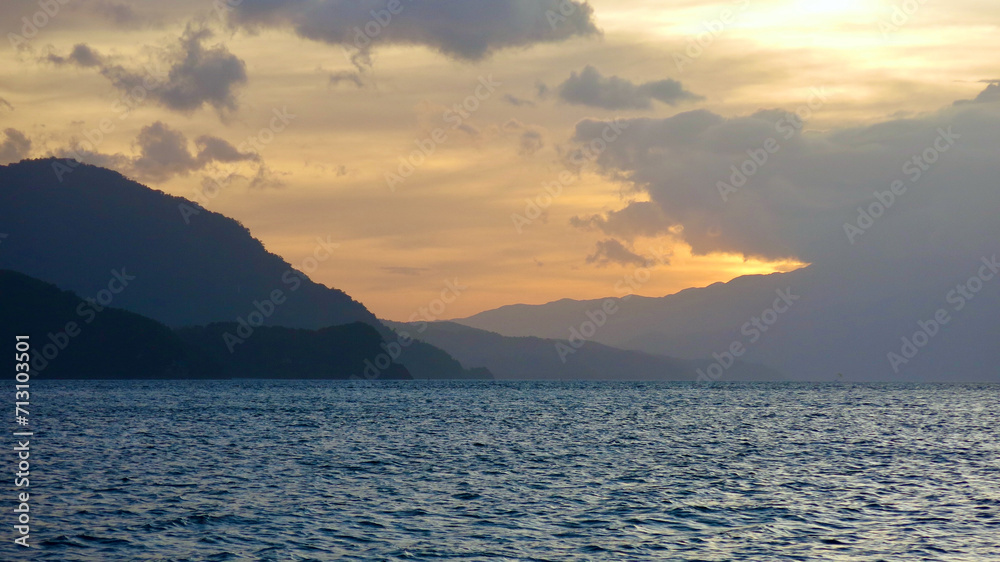 Tropical island at sunset. The sky is painted with bright colors by the rays of the setting sun over the ocean. Dark silhouette of a tropical island on the horizon during sunset. Tropical island at sunset. The sky is painted with bright colors by the rays of the setting sun over the ocean. Dark silhouette of a tropical island on the horizon during sunset.
