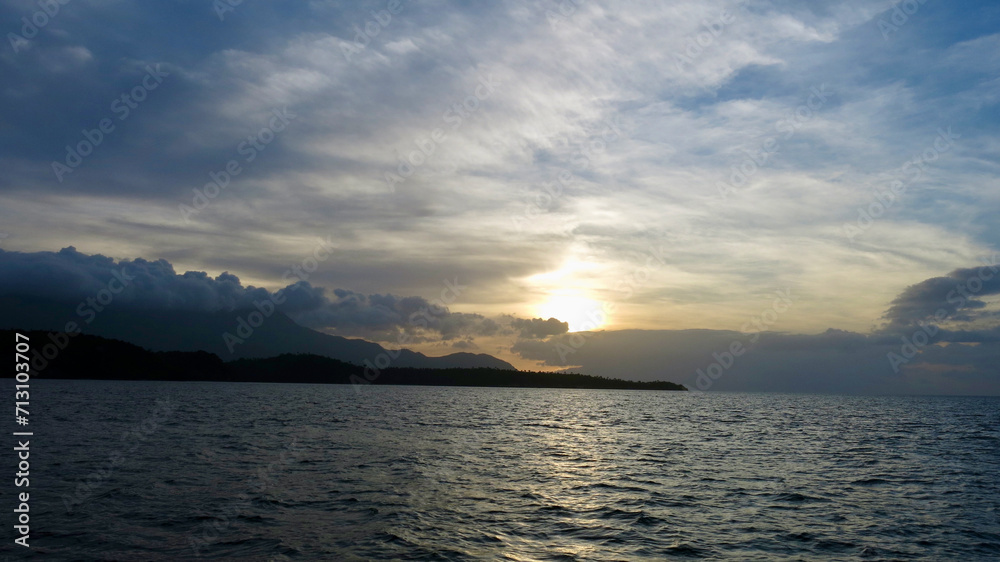 Tropical island at sunset. The sky is painted with bright colors by the rays of the setting sun over the ocean. Dark silhouette of a tropical island on the horizon during sunset.