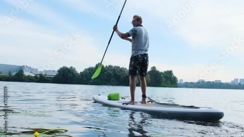Sportsman effortlessly navigates paddleboard with paddle. Man relishes active weekends spent on peaceful village lake with friends photo