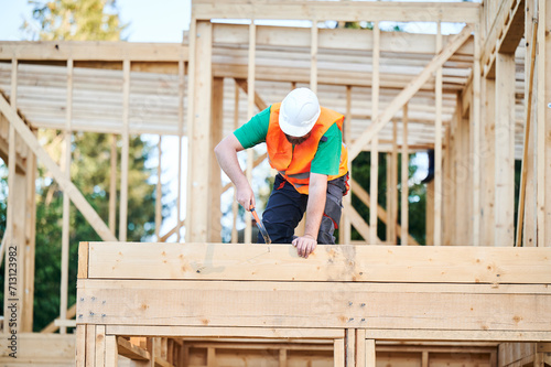 Carpenter constructing two-story wooden frame house. Bearded man hammering nails into structure while wearing protective helmet and construction vest. Concept of modern ecological construction.