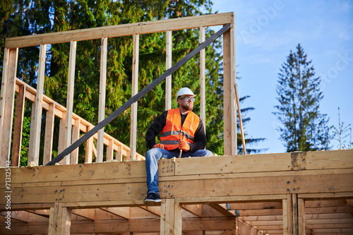 Carpenter constructing wooden frame two-story house near the forest. Bearded man holding hammer, dressed in protective helmet and orange safety vest. Concept of ecological modern construction.