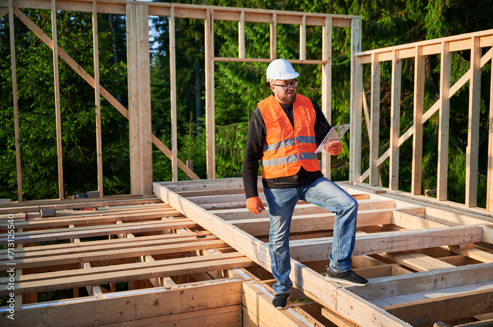 Engineer building wooden two-story structure near woods. Bearded with glasses man in jumpsuit and protective helmet examining construction plan. Concept of modern ecological construction. Engineer building wooden two-story structure near woods. Bearded with glasses man in jumpsuit and protective helmet examining construction plan. Concept of modern ecological construction.