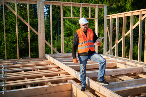 Engineer building wooden two-story structure near woods. Bearded with glasses man in jumpsuit and protective helmet examining construction plan. Concept of modern ecological construction.