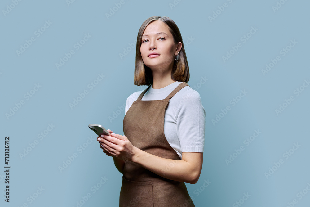 Portrait of young woman in apron holding smartphone in hands on blue background Portrait of young woman in apron holding smartphone in hands on blue background