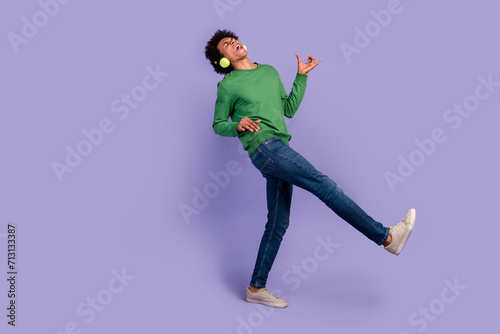 Full length photo of funky cheerful man dressed sweater earphones listening songs playing guitar empty space isolated violet color background
