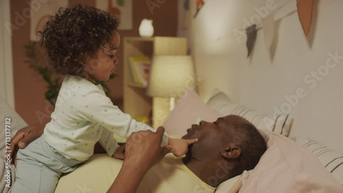 Medium side shot of African American father lying in bed and asking for kisses from little Biracial toddler girl who kissing him on mouth and cheeks in bedroom in eveningMedium side shot of African Am photo