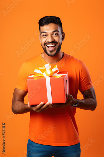 Young smiling happy Indian man wearing t-shirt casual clothes hold present box with gift ribbon bow looking aside on area isolated on orange red color background studio portrait. © AI_images