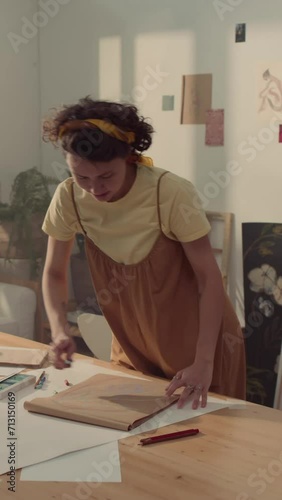 Medium full vertical shot of cheerful Caucasian female artist in jumpsuit standing by table in workshop, drawing sketch with pastel, then stepping away, looking at result and turning sheet aroundMediu photo