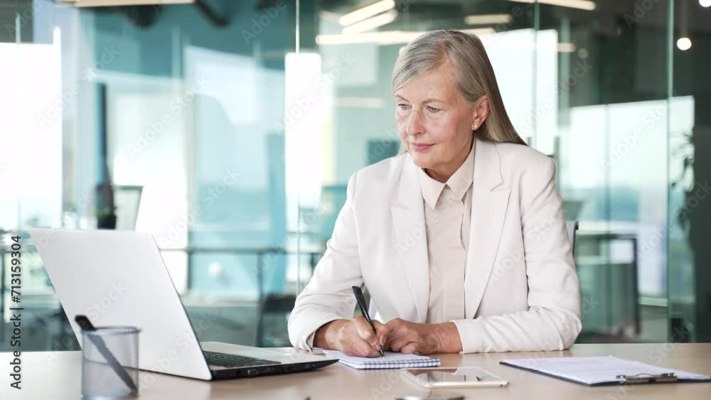 Senior gray haired businesswoman watching video call online conference taking notes looking at laptop computer screen sitting at workplace in office. Mature female listening remote business training Senior gray haired businesswoman watching video call online conference taking notes looking at laptop computer screen sitting at workplace in office. Mature female listening remote business training