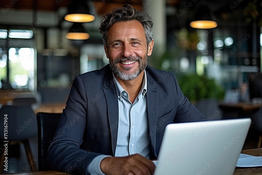 Middle aged businessman working at sunny work place on laptop while sitting at the wooden table. Blurred background. Middle aged businessman working at sunny work place on laptop while sitting at the wooden table. Blurred background.