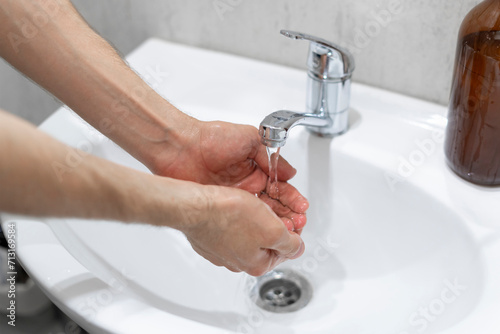 A close-up image capturing the act of handwashing, with a focus on hands scrubbing together under a running faucet over a white sink, next to a brown soap dispenser.